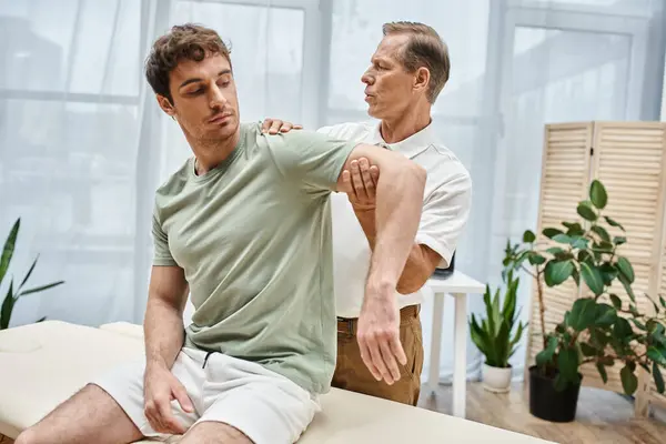 Handsome mature rehabilitologist in uniform helping patient to stretch during appointment in ward — Stock Photo