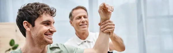 Jolly mature rehabilitologist helping merry patient to stretch on appointment in hospital, banner — Stock Photo
