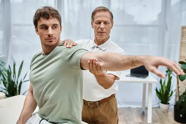 Attractive merry doctor in uniform helping his good looking patient to stretch, rehabilitation — Stock Photo