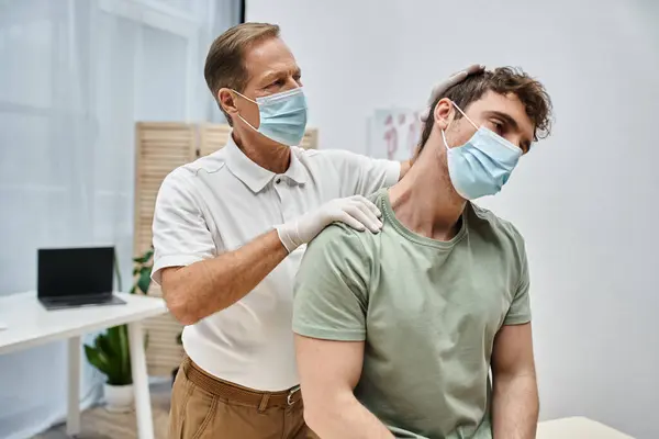 Rehabilitólogo dedicado con mascarilla y guantes masajeando el cuello de su paciente en la sala del hospital - foto de stock
