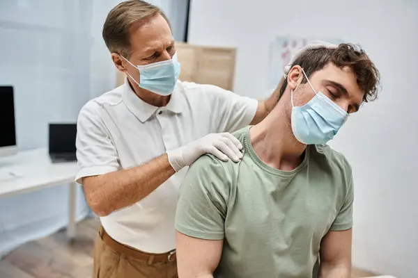 Dedicado médico maduro em uniforme com máscara massageando pescoço de seu paciente na enfermaria do hospital — Fotografia de Stock