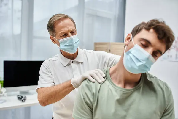 Dedicado rehabilitólogo maduro con mascarilla y guantes masajeando el cuello de su paciente en la sala del hospital - foto de stock