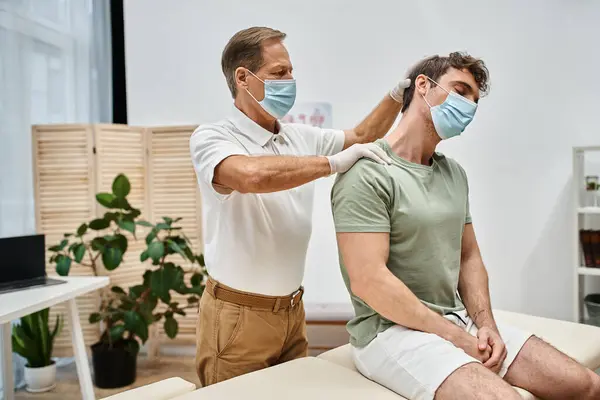 Devoted handsome mature doctor in uniform with mask massaging neck of his patient in hospital ward — Stock Photo