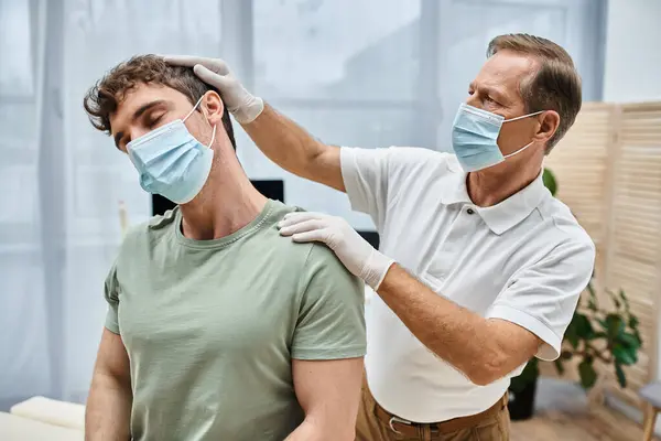 Médico maduro de buen aspecto con máscara y guantes ayudando a su paciente a rehabilitar en el hospital - foto de stock