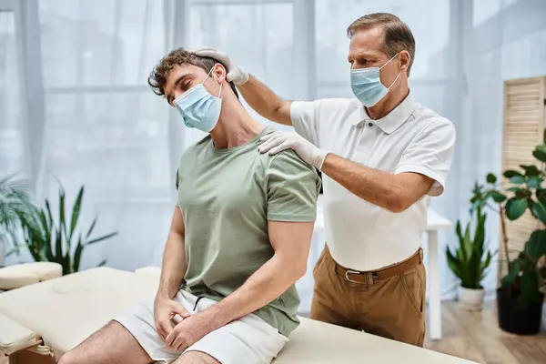 Guapo rehabilitólogo maduro con máscara y guantes masajeando el cuello de su paciente en la sala de hospital - foto de stock