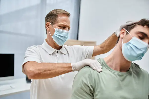 Médico maduro trabajador con máscara y guantes masajeando el cuello de su paciente en la sala del hospital - foto de stock
