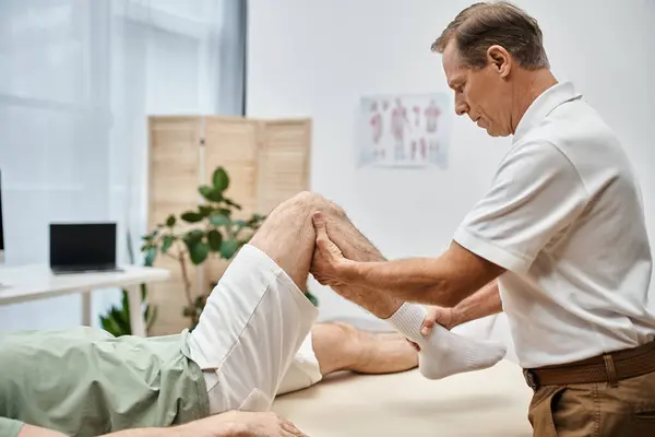 Bom olhar médico maduro verificando joelhos de seu paciente no hospital, conceito de reabilitação — Fotografia de Stock