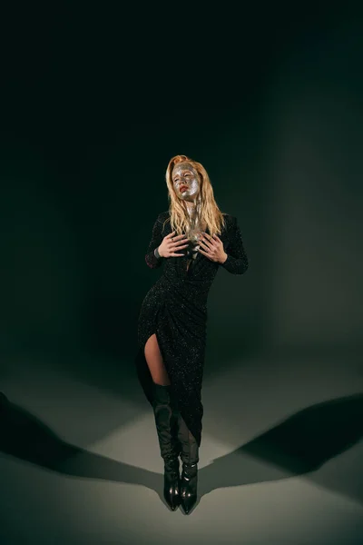Séduisante femme avec des paillettes sur le visage posant en robe brillante et genou bottes hautes sur fond noir — Photo de stock