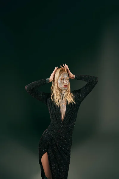 Блондинка з блиском на обличчі позує в блискучій сукні з руками біля волосся на чорному тлі — стокове фото