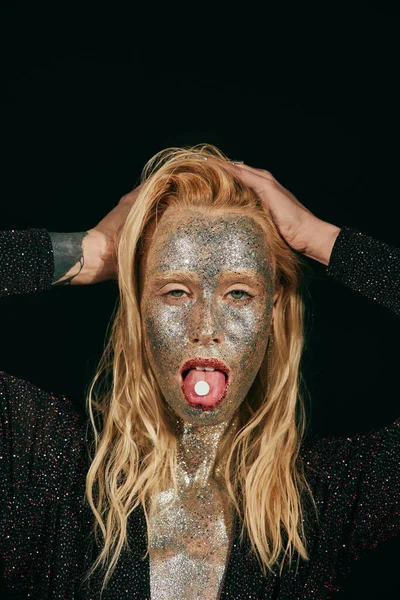 Provokante junge blonde Frau mit Glitzern im Gesicht, Zunge mit Pille auf schwarzem Hintergrund — Stockfoto