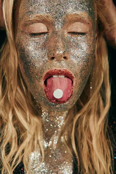 Close up of blonde woman with glitter makeup and closed eyes showing tongue with pill on black — Stock Photo