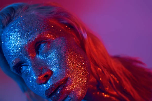 Nahaufnahme einer jungen und blonden Frau mit funkelndem Glitzern im Gesicht, die auf lebendigem Neonhintergrund posiert — Stockfoto