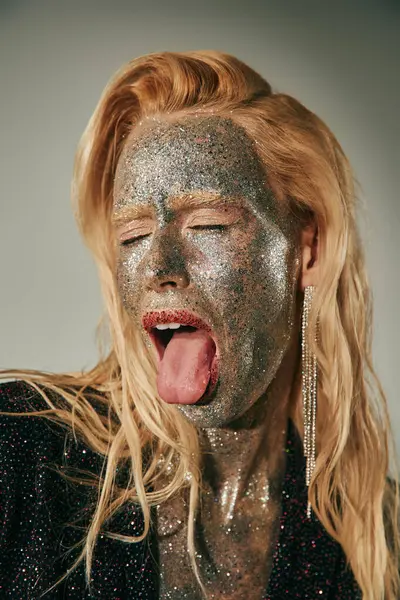 Provocative model with closed eyes and glitter on body and face sticking tongue on grey backdrop — Stock Photo