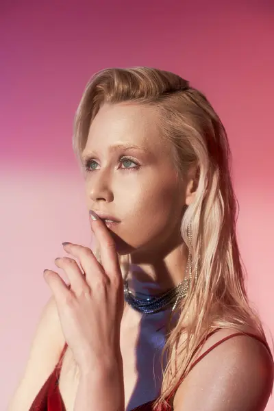 Задумчивая блондинка в роскошных украшениях позирует с пальцем рядом с губами на розовом фоне — стоковое фото