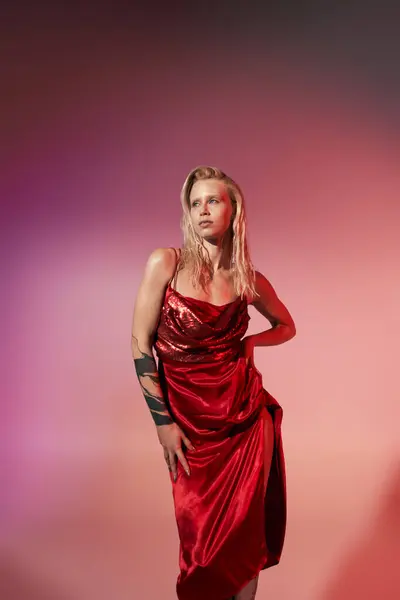 Bella giovane donna con tatuaggio in abito rosso in posa in movimento con mano sul fianco su sfondo rosa — Foto stock