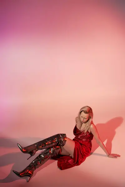 Appealing fashionable woman with long blonde hair in red dress sitting on floor on pink backdrop — Stock Photo