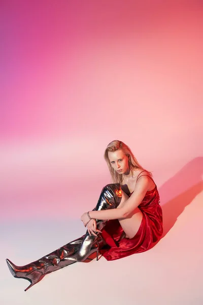 Beautiful stylish woman with long blonde hair in red dress sitting on floor on pink background — Stock Photo