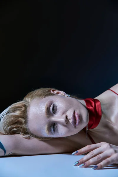 Модная блондинка с блестками на теле в красном элегантном платье лежа и глядя на камеру — стоковое фото