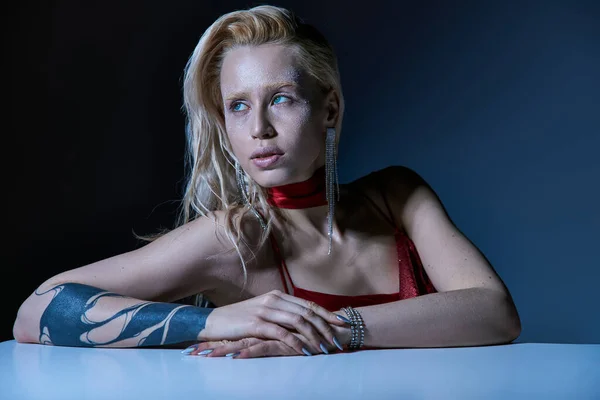 Fashionable young woman with highlighter and tattoo sitting and looking away on dark backdrop — Stock Photo