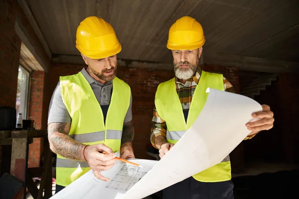 Dos constructores atractivos en cascos y chalecos de seguridad que trabajan con el plano antes de la construcción - foto de stock