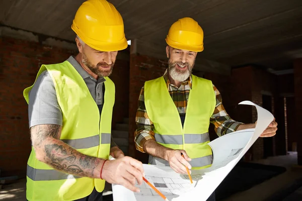 Cheerful good looking builders in safety vests and helmets looking at blueprint of building — Stock Photo