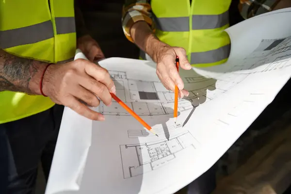 Ausgeschnittene Ansicht von zwei Bauarbeitern in Sicherheitswesten, die vor dem Bau mit dem Bauplan des Gebäudes arbeiten — Stockfoto