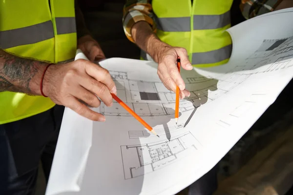 Vista ritagliata di due costruttori in giubbotti di sicurezza che lavorano con cianografia dell'edificio prima della costruzione — Foto stock