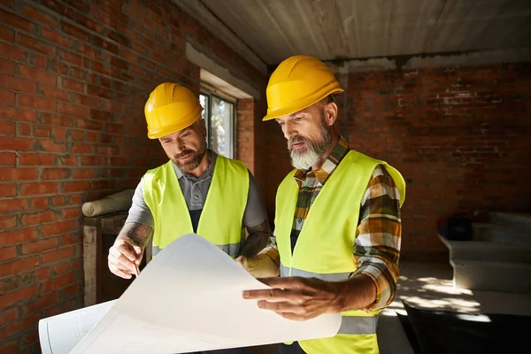 Two attractive builders in safety helmets and vests working with blueprint before construction — Stock Photo