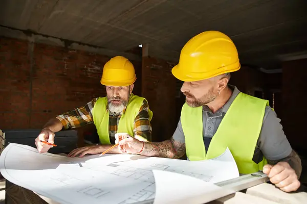 Dos constructores guapos en cascos de seguridad y chalecos que trabajan con planos antes de la construcción - foto de stock