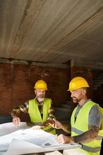 Two handsome builders in safety helmets and vests working with blueprint before construction — Stock Photo