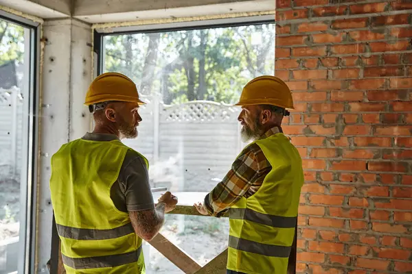 Two hardworking bearded men in safety vests and helmets on construction site, cottage builders — Stock Photo