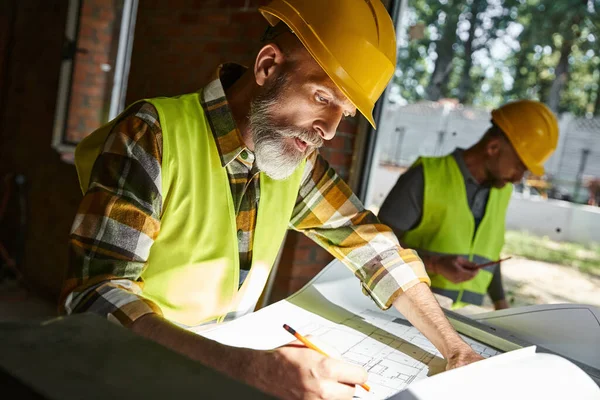Handsome worker looking at blueprint with his colleague working on backdrop, cottage builders — Stock Photo