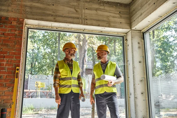 Hombres dedicados en chalecos de seguridad y cascos con planos en el sitio de construcción, constructores de casas de campo - foto de stock