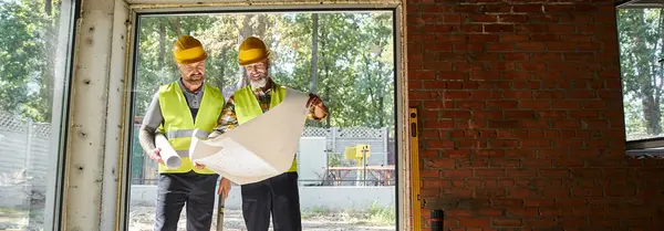 Hombres alegres en cascos de seguridad mirando el plano, mientras que en el sitio de construcción, constructores, pancarta - foto de stock