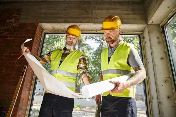 Trabajadores de la construcción guapos en cascos de seguridad que trabajan juntos en planos, constructores de casas de campo - foto de stock