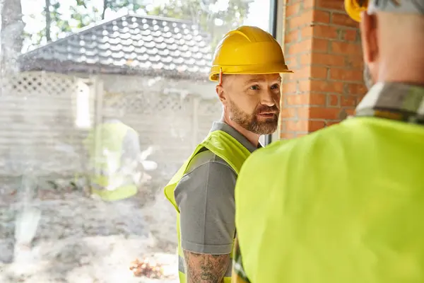 Enfoque en el trabajador barbudo en casco de seguridad hablando con su colega en el sitio de construcción, constructores - foto de stock