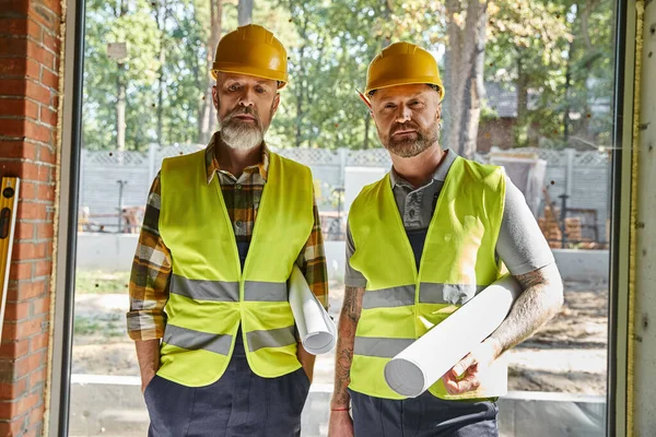 Good looking dedicated builders in safety vests and helmets looking at camera on construction site — Stock Photo