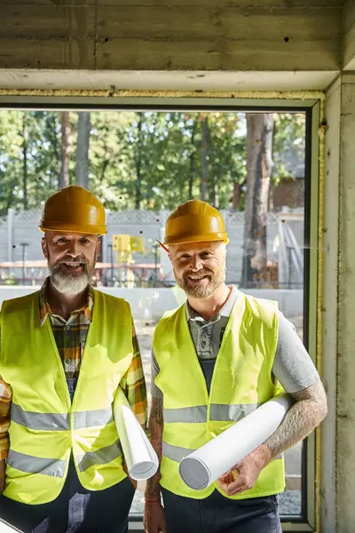 Cheerful dedicated builders in safety vests and helmets looking at camera on construction site — Stock Photo