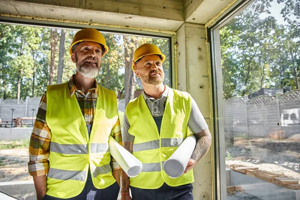 Trabajadores de la construcción alegres en chalecos de seguridad posando con planos y mirando hacia otro lado, constructores — Stock Photo