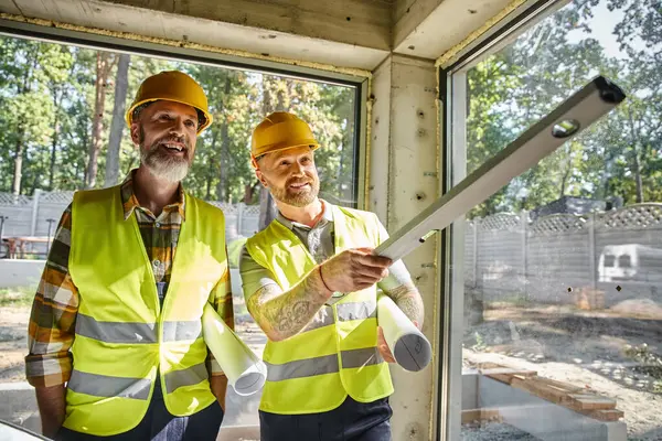Joyful construction workers in safety vests with blueprints and level looking away, builders — Stock Photo