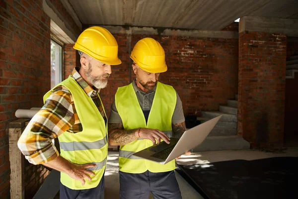 Good looking construction workers in safety vests working with laptop on site, cottage builders — Stock Photo