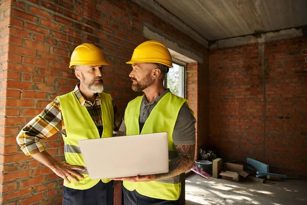 Hardworking construction workers in safety vests working with laptop on site, cottage builders — Stock Photo