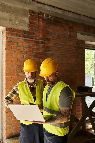 Hardworking construction workers in safety vests working with laptop on site, cottage builders — Stock Photo