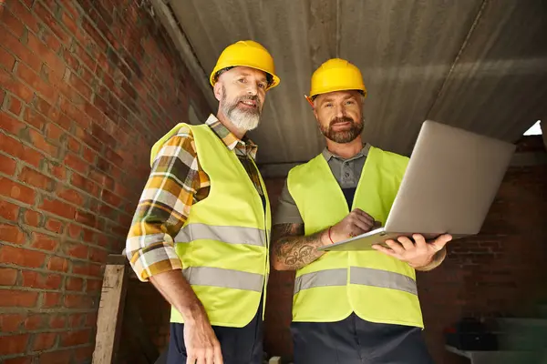 Trabalhadores de construção dedicados alegres em coletes de segurança que trabalham com laptop no local, construtores de casa de campo — Fotografia de Stock