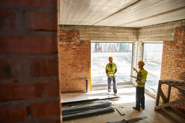 Dedicated appealing men in safety vests working on site with blueprints and level, cottage builders — Stock Photo