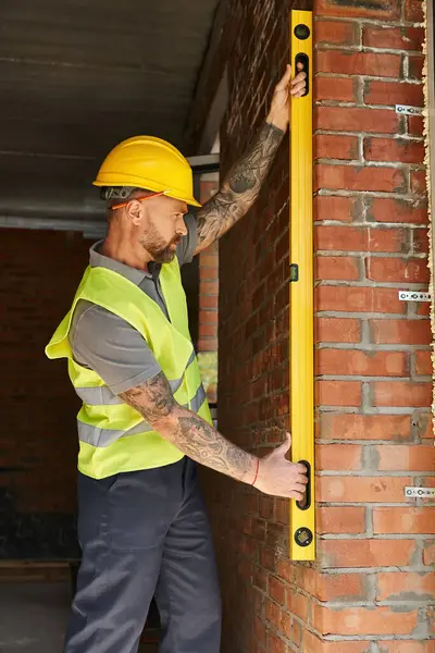 Trabalhador de construção barbudo bonito na parede de tijolo de medição de colete de segurança com nível, construtor — Fotografia de Stock