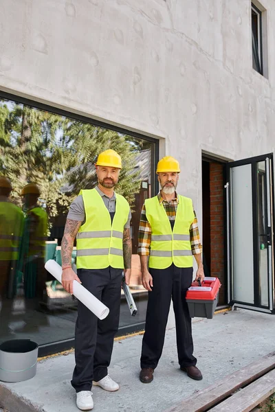 Trabajadores de la construcción alegres con nivel de caja de herramientas y plano mirando a la cámara mientras está fuera - foto de stock