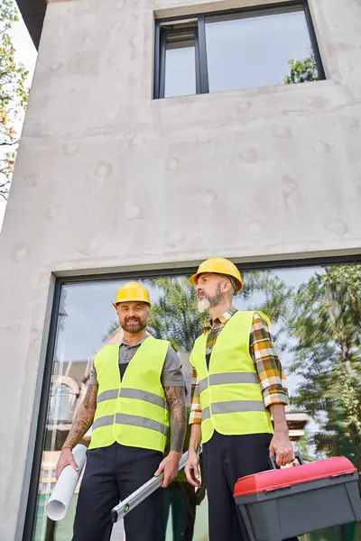 Trabajadores de la construcción alegres con nivel de caja de herramientas y plano mirando a la cámara mientras está fuera - foto de stock