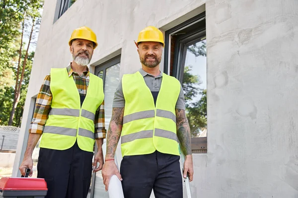 Frohe Bauarbeiter mit Werkzeugkiste und Blaupause, die draußen in die Kamera schauen — Stockfoto