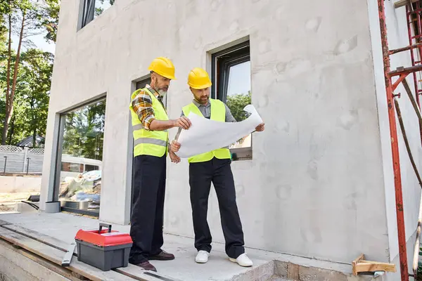 Good looking bearded builders in safety vests and helmets looking at blueprint while working outside — Stock Photo